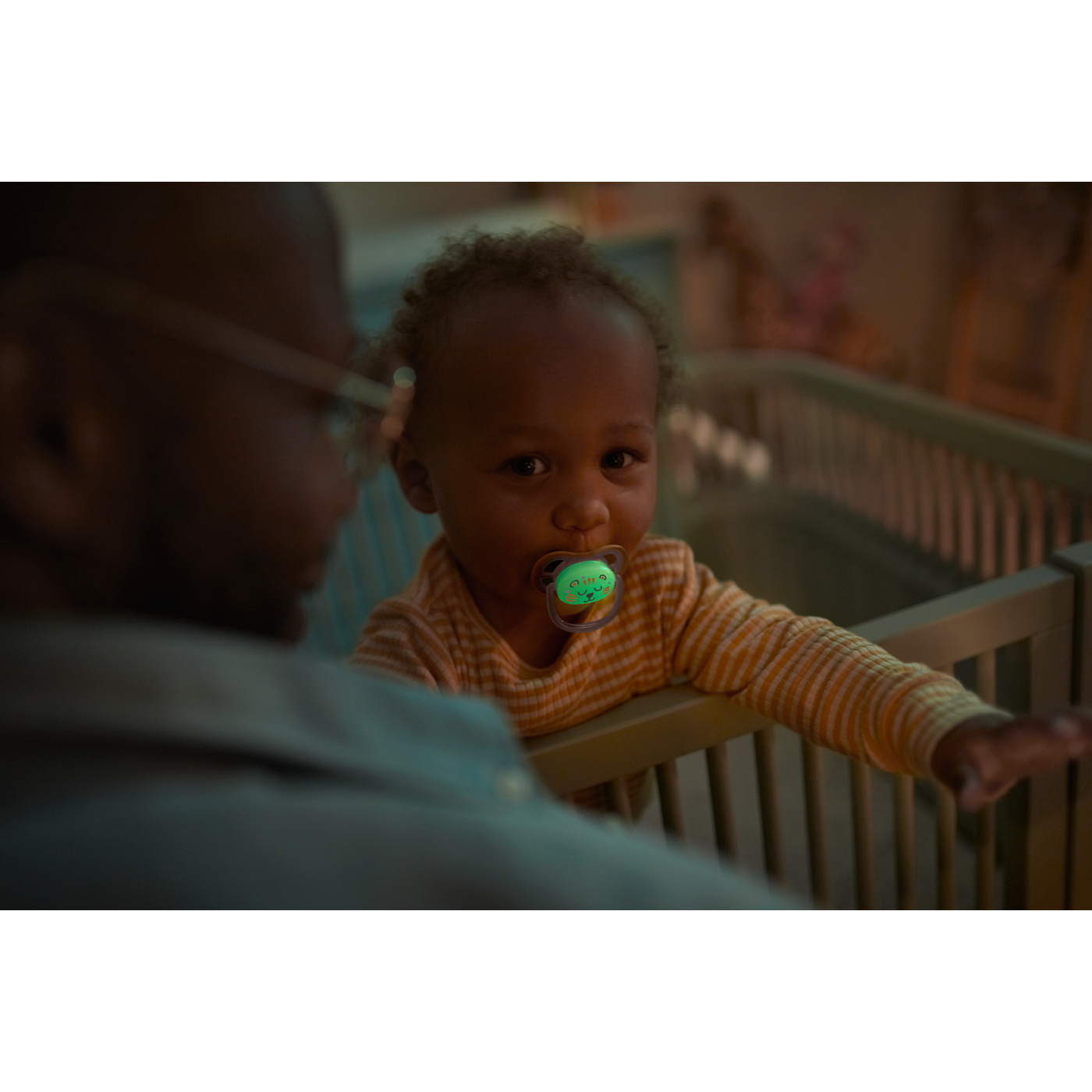 Baby in a crib with a pacifier, looking at an adult in a dimly lit room.