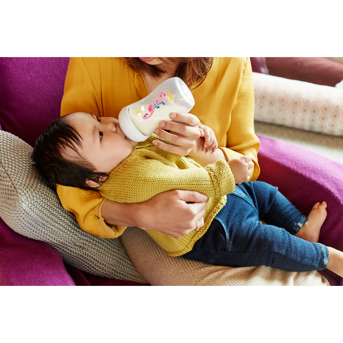 Person holding a baby and feeding them from a bottle on a couch.