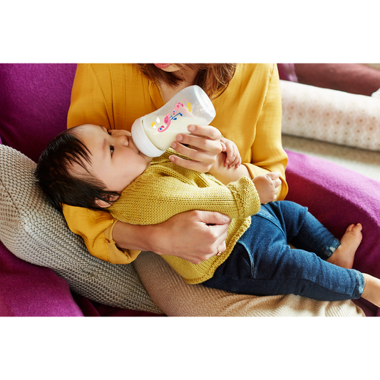 Person holding a baby and feeding them from a bottle on a couch.