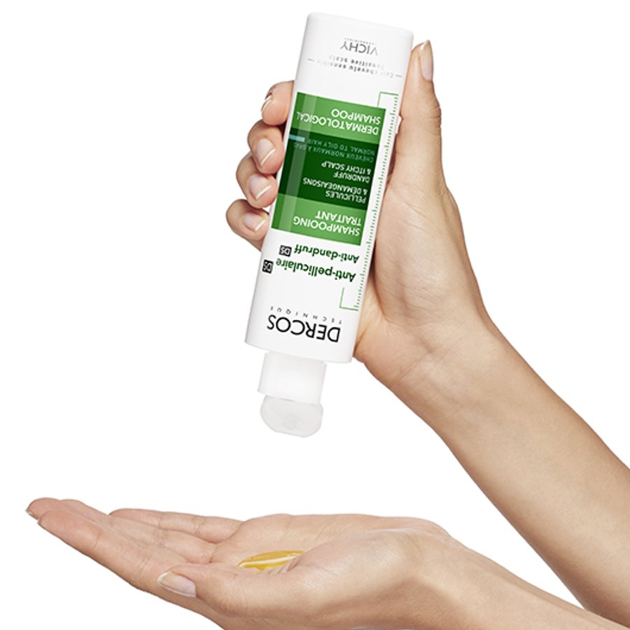 Hand holding a Dercos shampoo bottle with a small amount of product on the palm against a white background