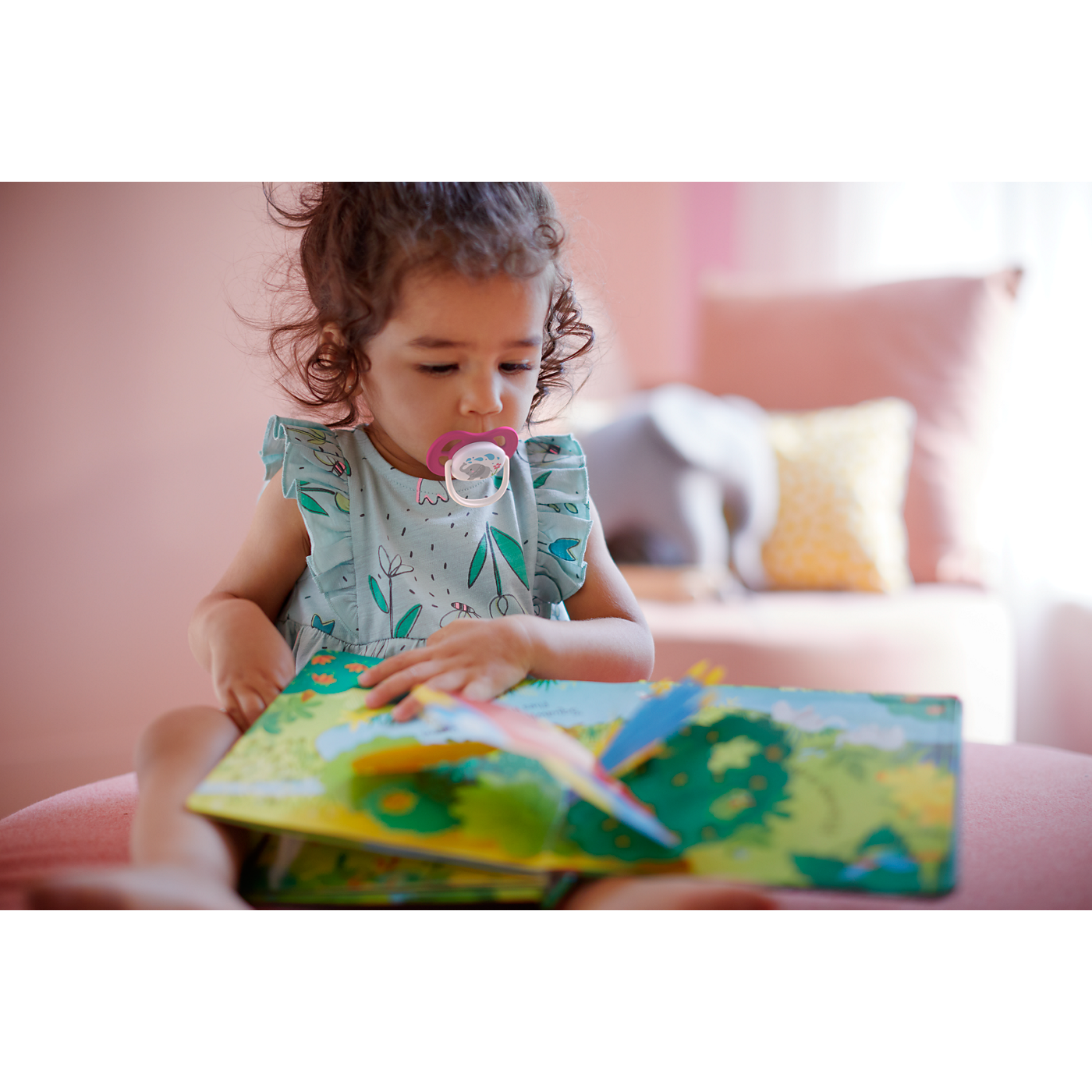 Child reading a book with a pacifier in their mouth, sitting on a couch.