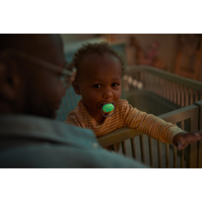 Baby in a crib with a pacifier, looking at an adult in a dimly lit room.