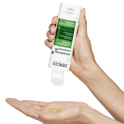 Hand holding a Dercos shampoo bottle with a small amount of product on the palm against a white background