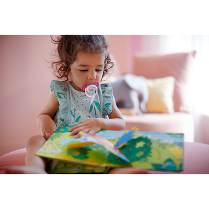 Child reading a book with a pacifier in their mouth, sitting on a couch.